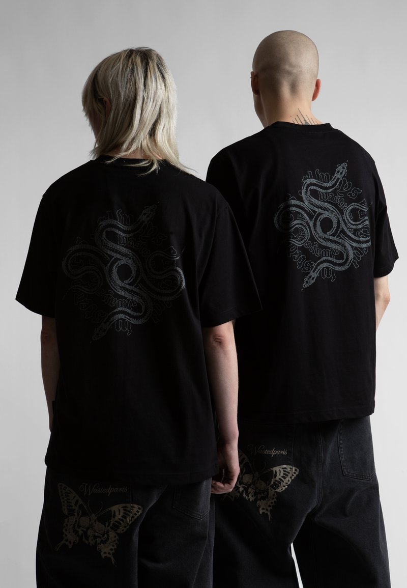 Két ember, akik fekete pólót és nadrágot viselnek, kígyó- és pillangómintával, hátukkal a kamera felé állnak.