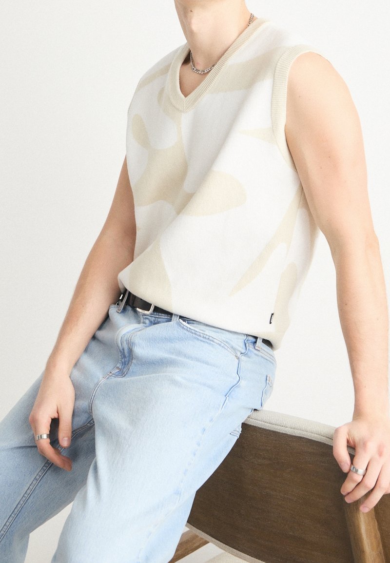 Personne portant un gilet sans manches beige et blanc à motifs, un jean bleu clair, un collier chaîne en argent et des bagues en argent, assise sur une chaise en bois.
