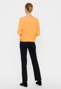 Oranges langärmliges Oberteil, kombiniert mit schwarzen geraden Jeans und Stiefeletten. Das Oberteil hat eine lockere Passform und einen offenen Ausschnitt.