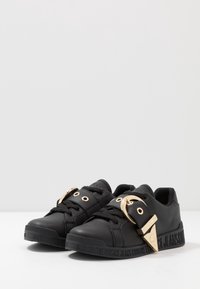 Baskets en cuir noir avec des accents en métal doré, comprenant une grande boucle et des œillets. Semelle texturée avec un logo latéral.