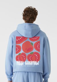 Šviesiai mėlyna treninginė palaidinė su dideliu raudonų pomidorų grafikos piešiniu ant nugaros, kuriame yra užrašas "PIZZA ESSENTIALS" balta spalva. Minkštas audinys, kasdieniškas dizainas.