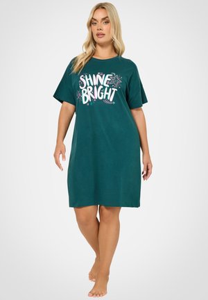 Vrouw in een teal knielange jurk met de tekst "SHINE BRIGHT" en stergrafieken, die blootsvoets staat tegen een effen witte achtergrond.