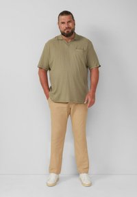 Olijfgroene polo met korte mouwen en pocket, gecombineerd met beige lichte broeken en witte sneakers in een staande houding.