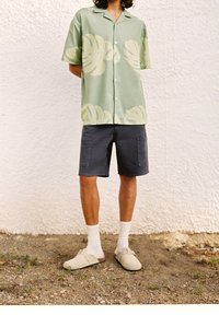 Grünes Kurzarmhemd mit hellem Blattmuster, graue Shorts mit Taschen, weiße Knöchelsocken und beige Slip-On-Schuhe, stehend auf Kies.