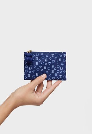 Una mano sostiene una bolsa con cremallera azul marino con patrones de palos de cartas en azul claro y un tirador de cremallera con pompón azul marino sobre un fondo liso.