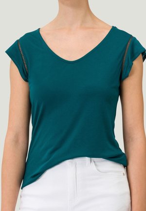 Turquoise top met korte mouwen en V-hals met doorschijnende mesh strookdetails op de mouwen, gedragen ingestopt in een witte broek.