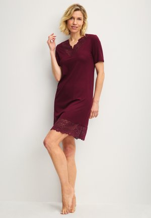Donna bionda con vestito bordeaux a maniche corte con rifiniture in pizzo sullo scollo e sull'orlo, in piedi a piedi nudi contro un muro bianco.