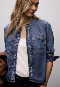 Femme aux longs cheveux blonds portant une veste boutonnée en denim bleu par-dessus un haut blanc, tenant un vêtement marron dans sa main gauche.