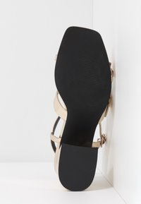 Sandale à talon haut beige avec semelle noire, présentant plusieurs fines lanières et une finition lisse. Détail boucle sur le côté extérieur.