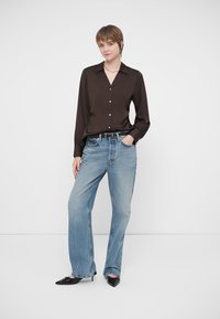 Noor inimene seisab ühe käega taskus, seljas must nööpideta särk, heledad sinised teksad, must vöö ja mustad teravapealised kingad.