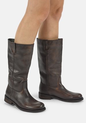 Gambe con stivali alti in pelle marrone scuro con tacchi bassi impilati, in piedi su una superficie chiara con uno sfondo semplice.