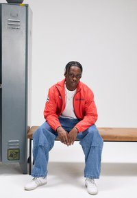 Röd vindjacka med texten "REVOLUTION", vit t-shirt, lösa blå jeans, sittande på en träbänk nära ett grått skåp. Vita sneakers.