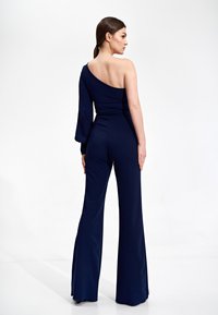 Granatowy kombinezon z jedną długą rękawą i jednym odkrytym ramieniem, dopasowany gorset i rozszerzane nogawki, wykonany z gładkiego, elastycznego materiału.
