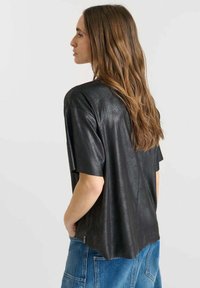 Donna con lunghi capelli castani che indossa una maglietta nera a maniche corte larga e pantaloni in denim blu, rivolta a sinistra con le mani nelle tasche.
