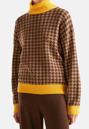 Pull à motif pied-de-poule marron et beige avec col roulé, poignets et ourlet côtelés jaunes, porté avec un pantalon marron.