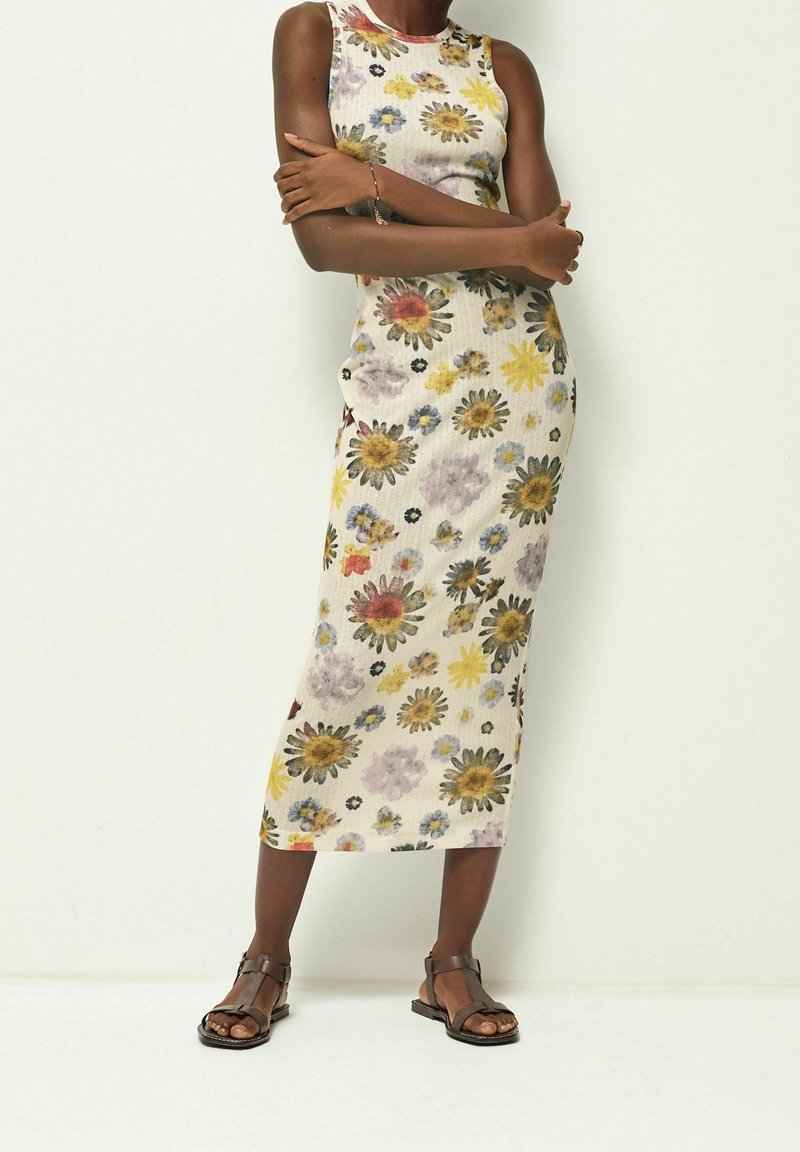 Robe longue fleurie à coupe ajustée, ornée de fleurs jaunes et violettes sur un fond clair. Portée avec des sandales marron à lanières.