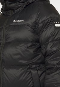 Veste Columbia noire matelassée avec fermeture éclair à l'avant, logo sur la poitrine et écusson de marque sur le haut de la manche.