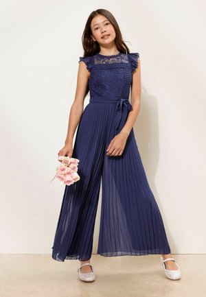 Pige iført en marineblå blonde- og plisseret jumpsuit, der holder en buket lyserøde roser og står mod en lys baggrund.