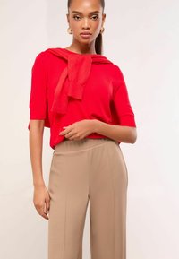 Red short-sleeve top with a knotted detail at the neckline, paired with beige high-waisted trousers featuring vertical seam lines.