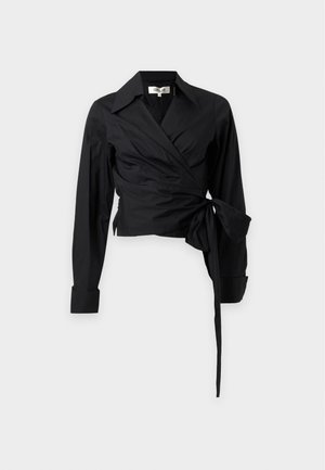 Femme debout, les mains sur les hanches, portant un chemisier cache-cœur noir et un pantalon large avec un imprimé zèbre noir, blanc et bleu, ainsi que des sandales noires.