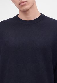 Gros plan d'une personne portant un pull uni bleu marine foncé à col rond côtelé.