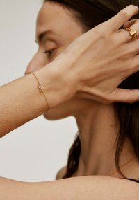 Pulsera de oro en la muñeca con un diseño de clip de papel. La cadena es delgada con un acabado texturizado, realzando su apariencia delicada.