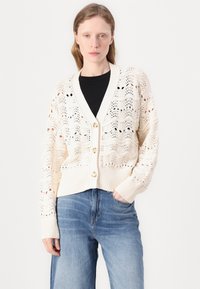 Strikkegenser i elfenben med et blomstermønster i åpen veving, med dyp V-hals, lange ermer og store kremfargede knapper.