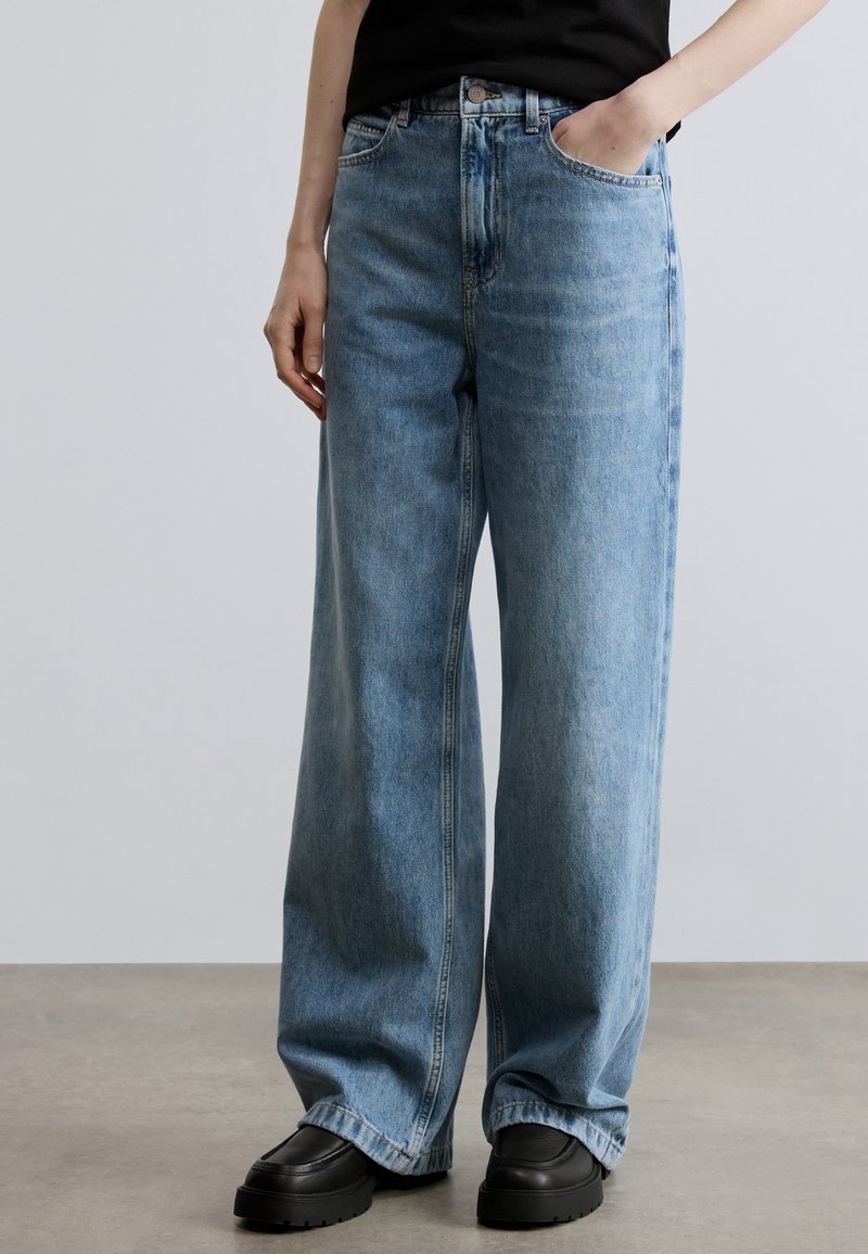 Jean en denim à jambes larges de couleur bleu clair, avec une taille haute, deux poches avant, et une texture légèrement délavée. Porté avec des chaussures noires épaisses.