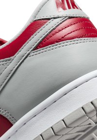 Sneaker in pelle grigia con accenti rossi, superficie texture, colletto imbottito e suola in gomma bianca con un design del battistrada a motivo.