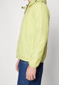 Jasnozielona kurtka przeciwwietrzna z zamkiem błyskawicznym z przodu, regulowanym kapturem i bocznymi kieszeniami. Gładka faktura i elastyczne mankiety.
