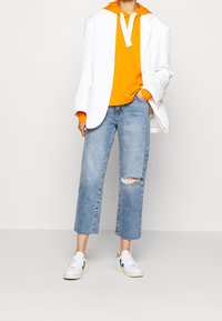 Blazer bianco, felpa arancione con cappuccio, jeans chiari strappati e sneakers bianche con dettagli neri. Outfit casual a strati con un mix di texture.