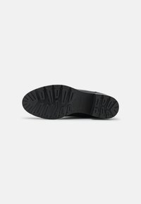 Semelle de chaussure noire en caoutchouc texturé, avec des rainures et des creux profonds pour une meilleure adhérence. Forme plate et arrondie, avec une finition globale lisse.