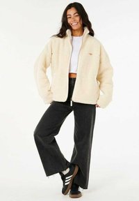 Crème-kleurige fleece jas met een ritsluiting, hoge kraag en twee voorzakken, gecombineerd met zwarte wijde jeans en zwarte sneakers.