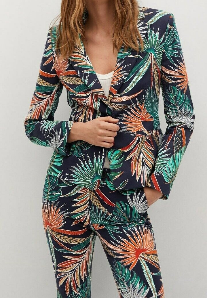 Femme portant un blazer et un pantalon sombres avec un motif coloré de feuilles tropicales, debout, une main dans la poche et l'autre ajustant le blazer.