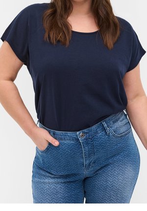 Kobieta ubrana w granatową koszulkę z krótkim rękawem wpuszczoną do wysokowasowych niebieskich dżinsów z wzorem, z jedną ręką w kieszeni.