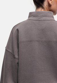 Felpa grigia oversized con colletto alto, cuciture in vista e una texture morbida, mostrata da dietro. Design minimalista.