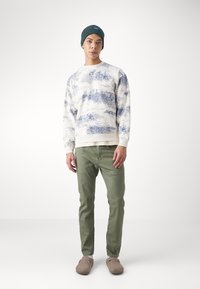 Lys kremfarget genser med blått landskapsmotiv, grønne slim-fit bukser, grå slip-on sko og en mørkegrønn beanie.