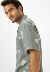 Kurzärmliges Hemd aus hellgrünem Stoff mit einem floralen Muster in Weiß und hellem Gelb. Mit einer Brusttasche und einer lockeren Passform.
