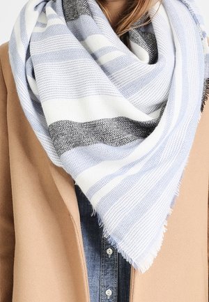 Femme portant une grande écharpe en tricot rayée blanche, bleu clair et noire sur un manteau camel et une chemise en denim.