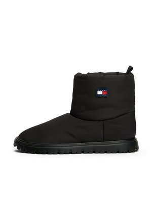 Bottine noire matelassée avec un bout rond et une semelle crantée. Elle présente une étiquette de logo sur le côté extérieur et une boucle de traction en haut.