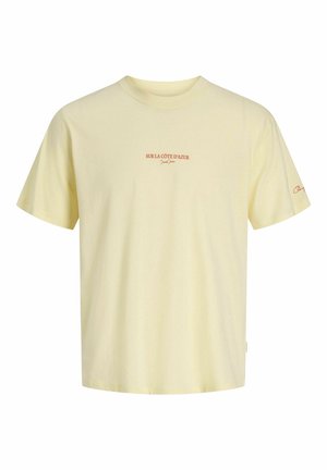 Ung person iført en lysegul T-shirt, mørke slidte denimshorts, beige sneakers, hvide sokker og en pink baseballkasket, står og kigger mod højre.