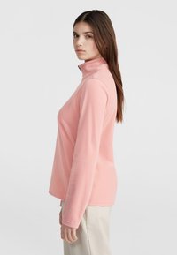 Light pink fleece top with a high collar and long sleeves, featuring smooth texture and a relaxed fit. No visible logos or patterns.