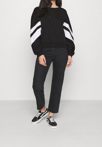 Sudadera negra con rayas blancas en las mangas, cuello redondeado, corte relajado, combinada con jeans negros de pierna recta y zapatillas negras.