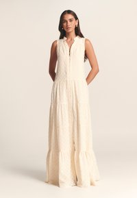 Maksik obleka v kremni barvi z teksturiranim vzorcem, brez rokavov, z gumbi spredaj in večnivojsko krilom. Opremljena je s ovratnikom in stranskimi žepi.