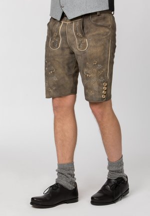 Mann trägt traditionelle braune Lederhosen mit Stickerei, graue Wollsocken, schwarze Schuhe und eine graue Weste.