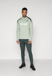 Sportswear sæt med en lys grøn langærmet top med lynlås krave og mørke grønne tætsiddende bukser, accentueret med logodetaljer og sort fodtøj.