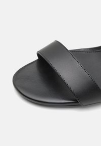 Sandal in pelle nera con una fascia larga, superficie liscia, punta arrotondata e suola piatta. Il design enfatizza il minimalismo e la funzionalità.