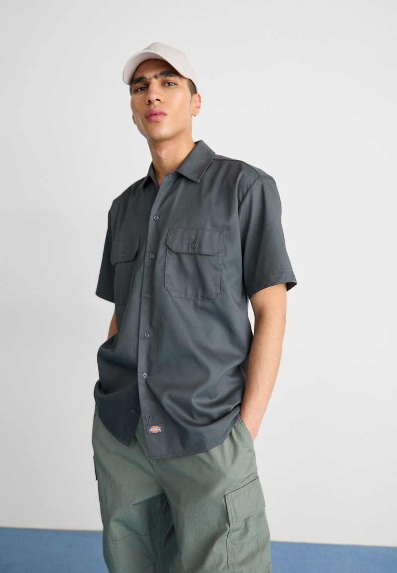 Camicia grigia a maniche corte con bottoni, due tasche sul petto e un piccolo segno del logo. Abbinata a pantaloni cargo verde chiaro.