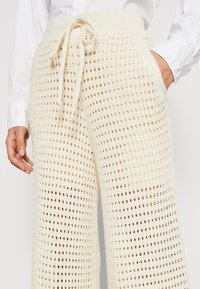 Luźne spodnie wykonane na szydełku, w kremowym kolorze, z szerokim pasem z troczkiem oraz teksturowanym, ażurowym wzorem na całej powierzchni.