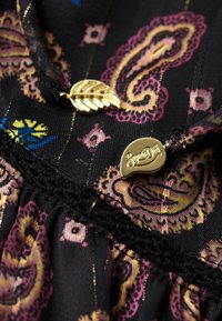 Tissu noir avec motif paisley rose, bordure en dentelle noire et deux boutons en métal doré, l'un en forme de feuille et l'autre gravé avec "SuperDry".